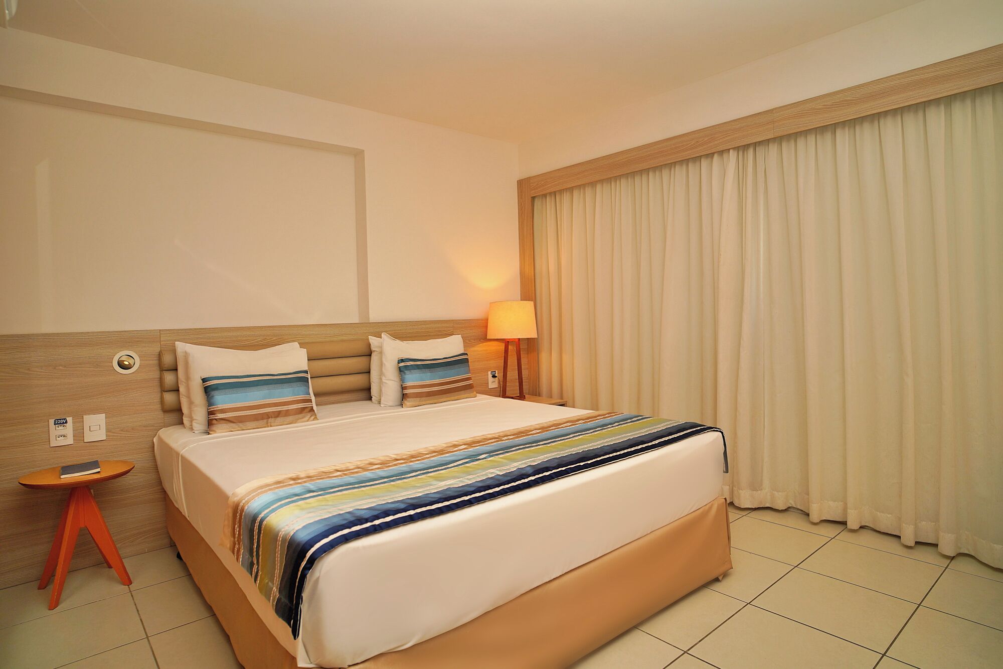Quarto de hotel com uma cama grande, mesa de cabeceira, abajur e janela com cortinas.