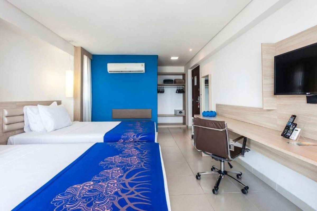 A hotel room with a bed, a desk, a chair, a TV, and a blue wall.
