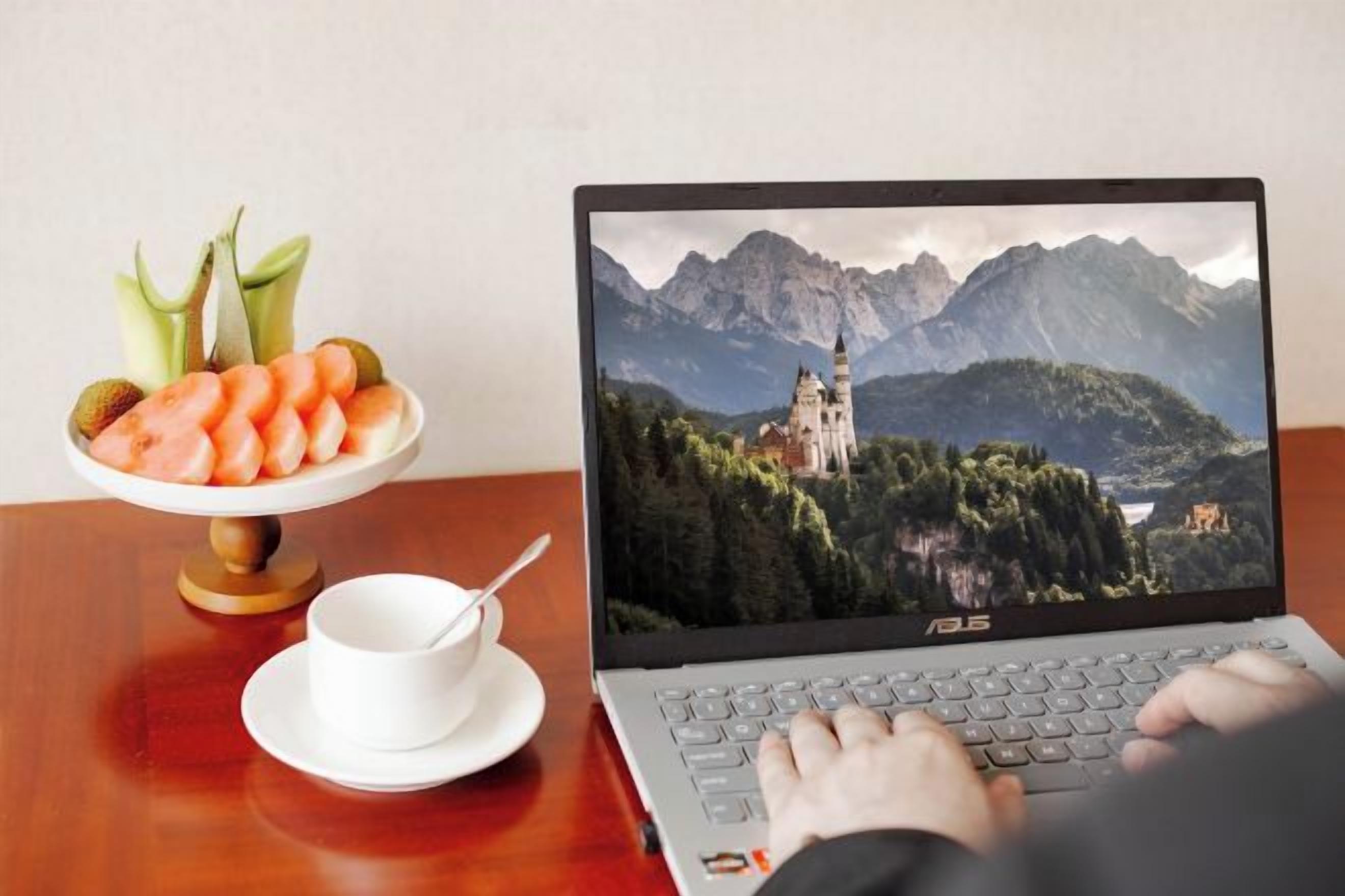A laptop on a wooden table with a scenic mountain view on the screen, a bowl of fruit, and a white cup with a spoon.
