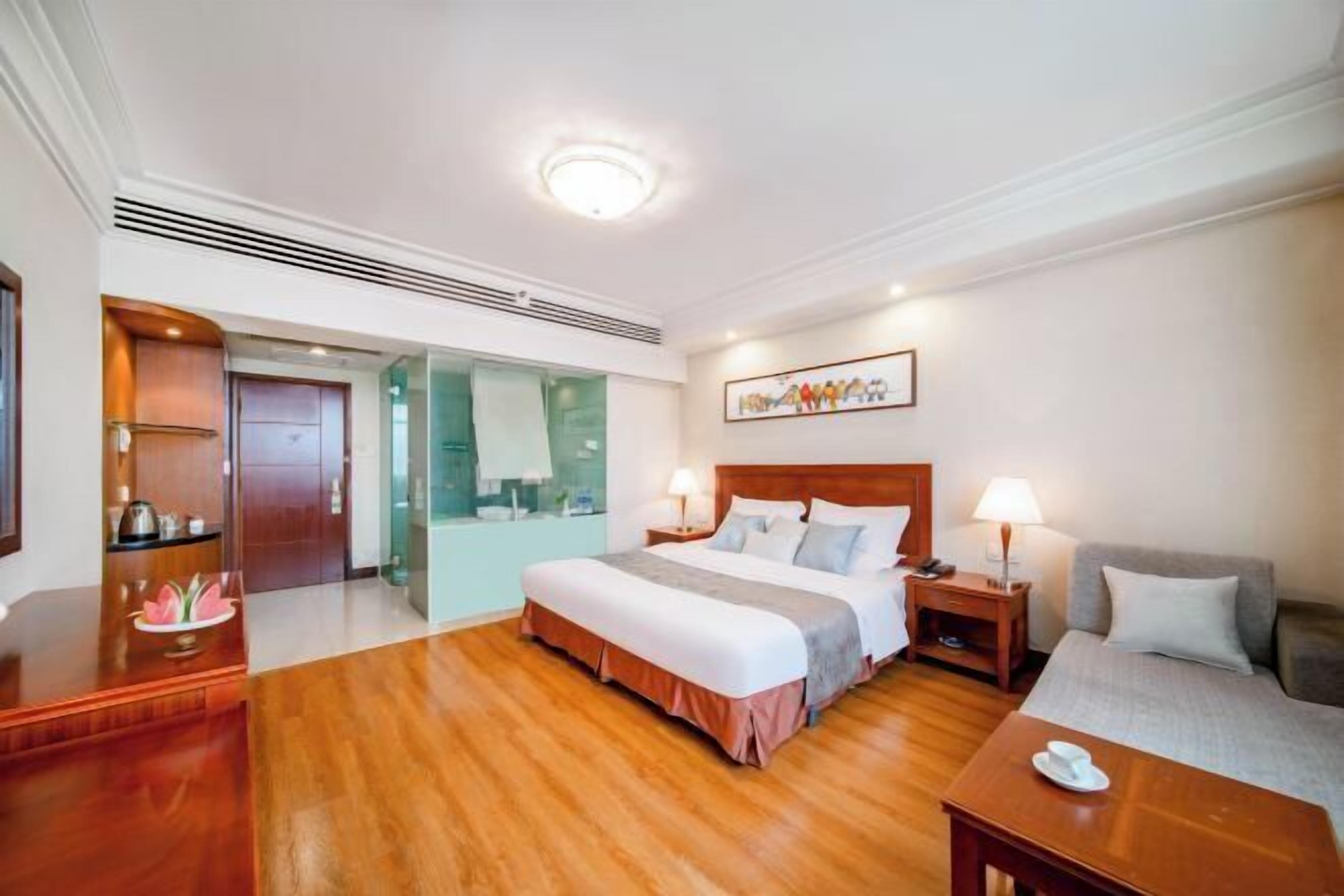 A hotel room with a wooden floor, a bed with a wooden headboard, a bedside table with a lamp, a sofa, and a desk with a lamp.