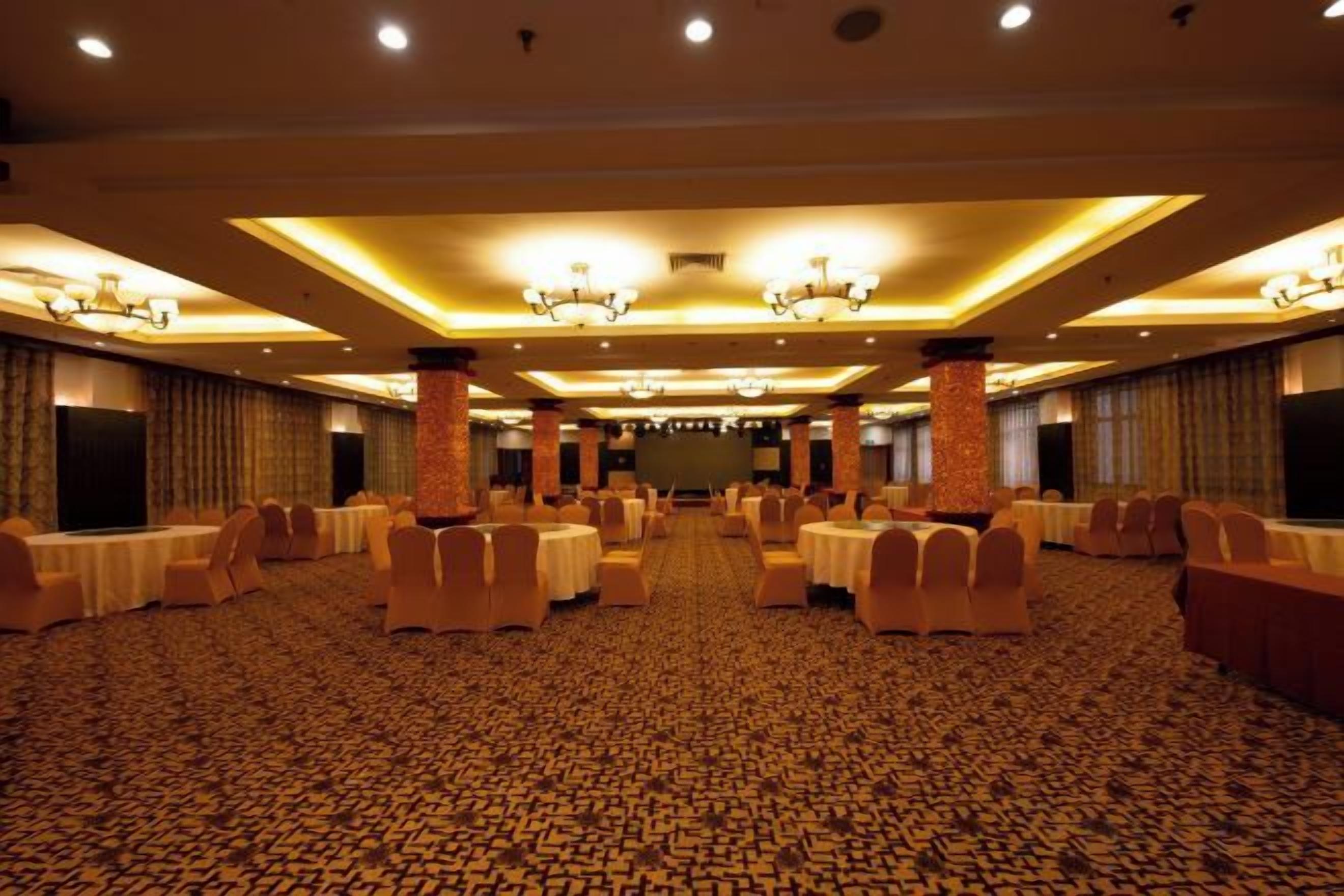 A large banquet hall with round tables and chairs, high ceilings with chandeliers, and a patterned carpet.