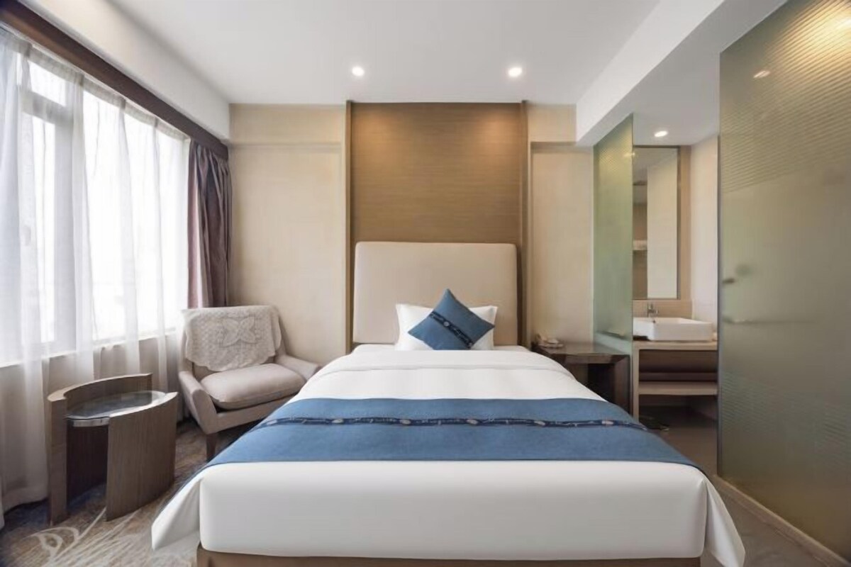 Habitación de hotel con una cama grande, una silla, una mesita de noche, un espejo y un baño con lavamanos y espejo.