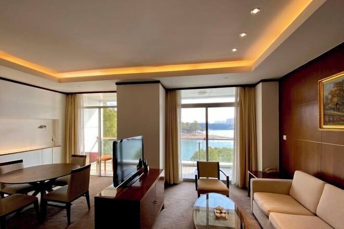 Habitación de hotel con comedor, sofá, televisor y un amplio ventanal con vistas al exterior.