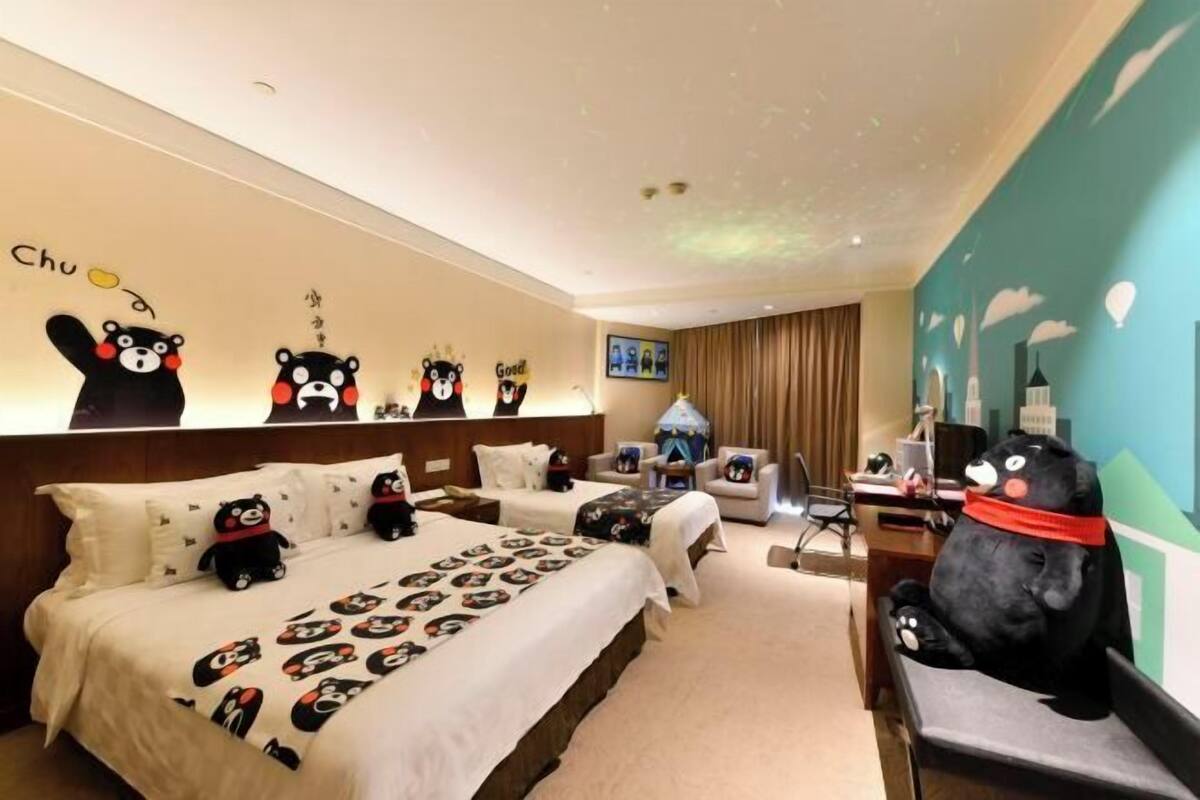 Una habitación de hotel con una cama grande, dos mesitas de noche, un escritorio y una silla. Hay decoraciones en la pared y un proyector en el techo.