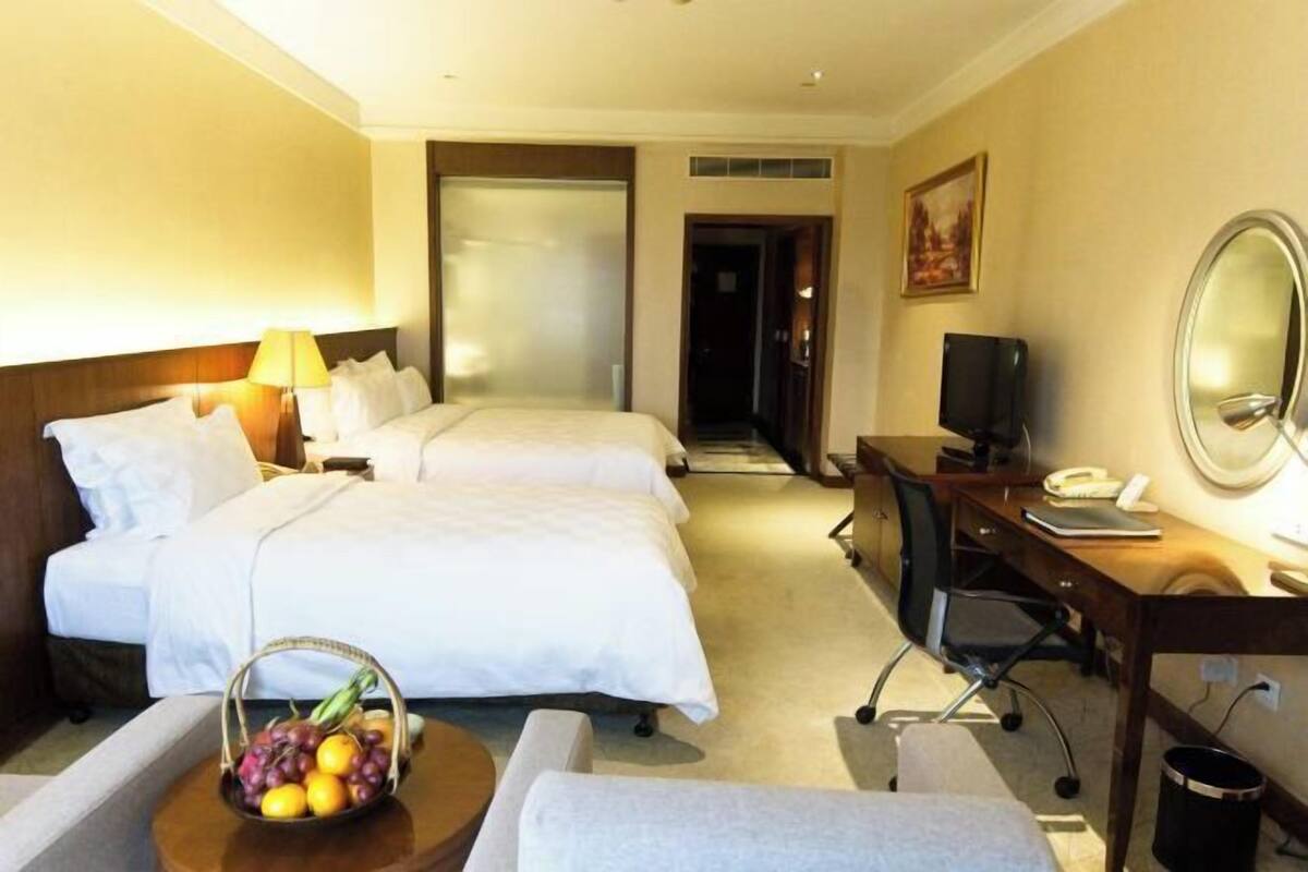 Habitación de hotel con dos camas, un escritorio, una silla, un televisor, una lámpara de escritorio, un teléfono y una cesta de frutas.