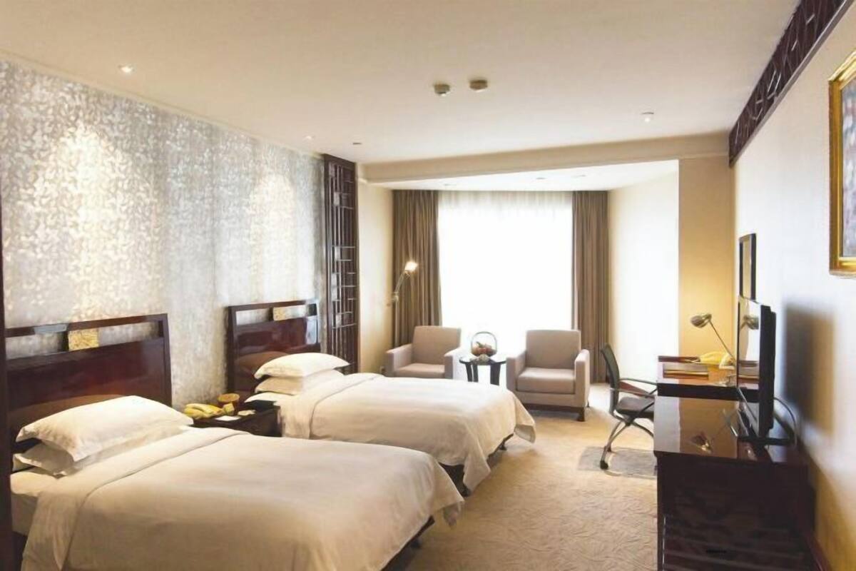 Habitación de hotel con dos camas, un escritorio, una silla, un televisor y un ventanal con cortinas.