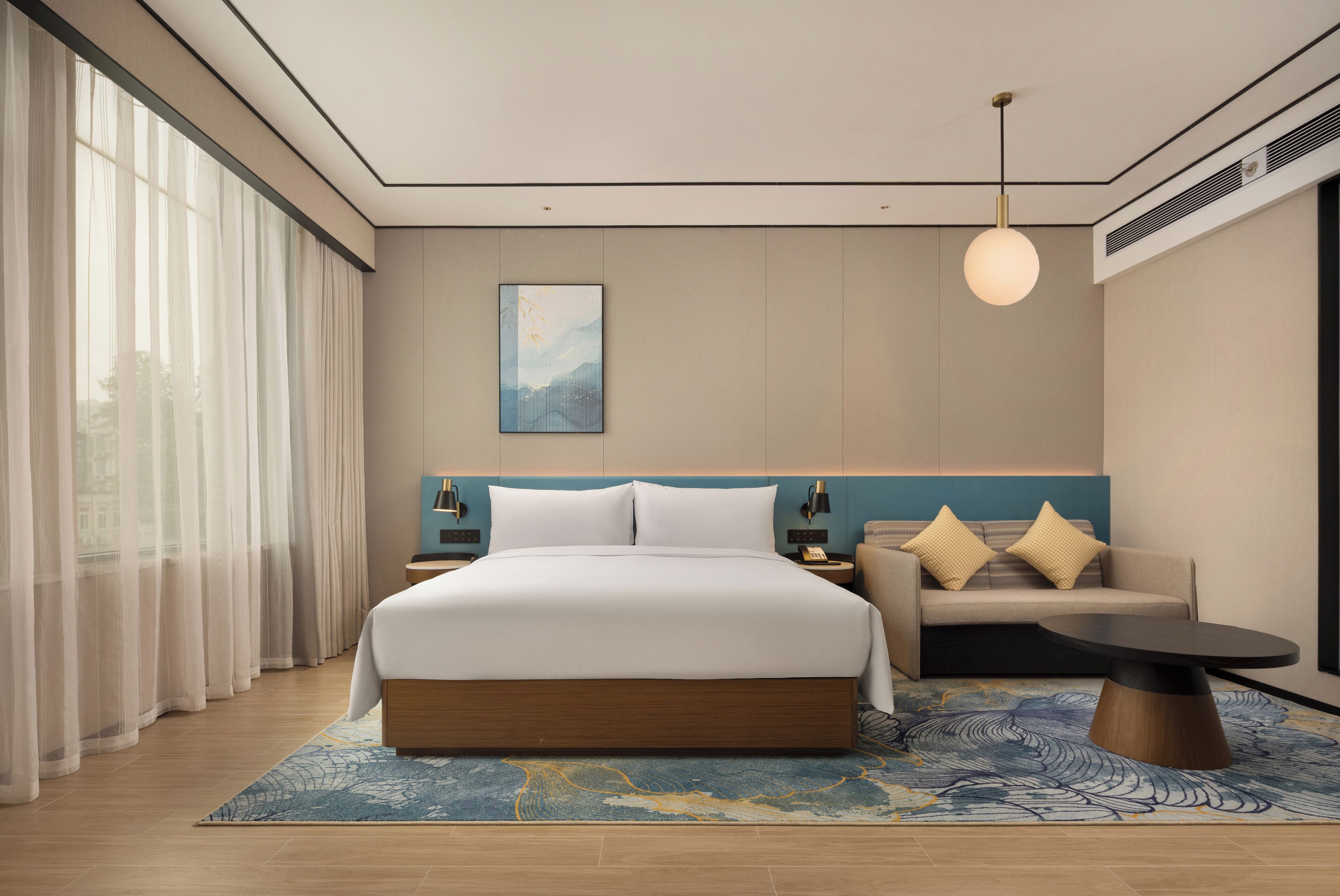 A modern hotel room with a large bed, a sofa, a coffee table, and a view of the city through sheer curtains.
