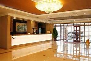 Ein Hotel-Empfangsbereich mit Marmorboden, einem großen Kronleuchter, einer Rezeption, einem gerahmten Kunstwerk und einem großen Fenster mit Außenblick.