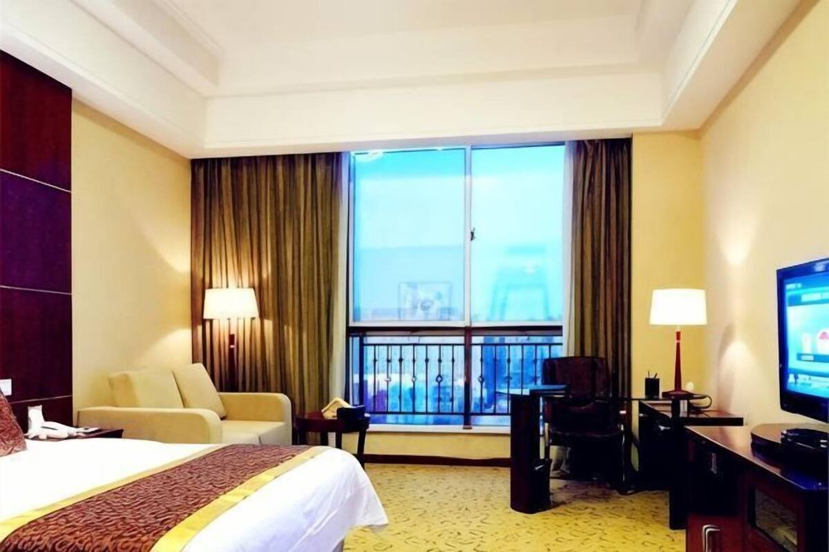 A hotel room with a bed, two chairs, a desk with a television, a desk lamp, and a large window with curtains.