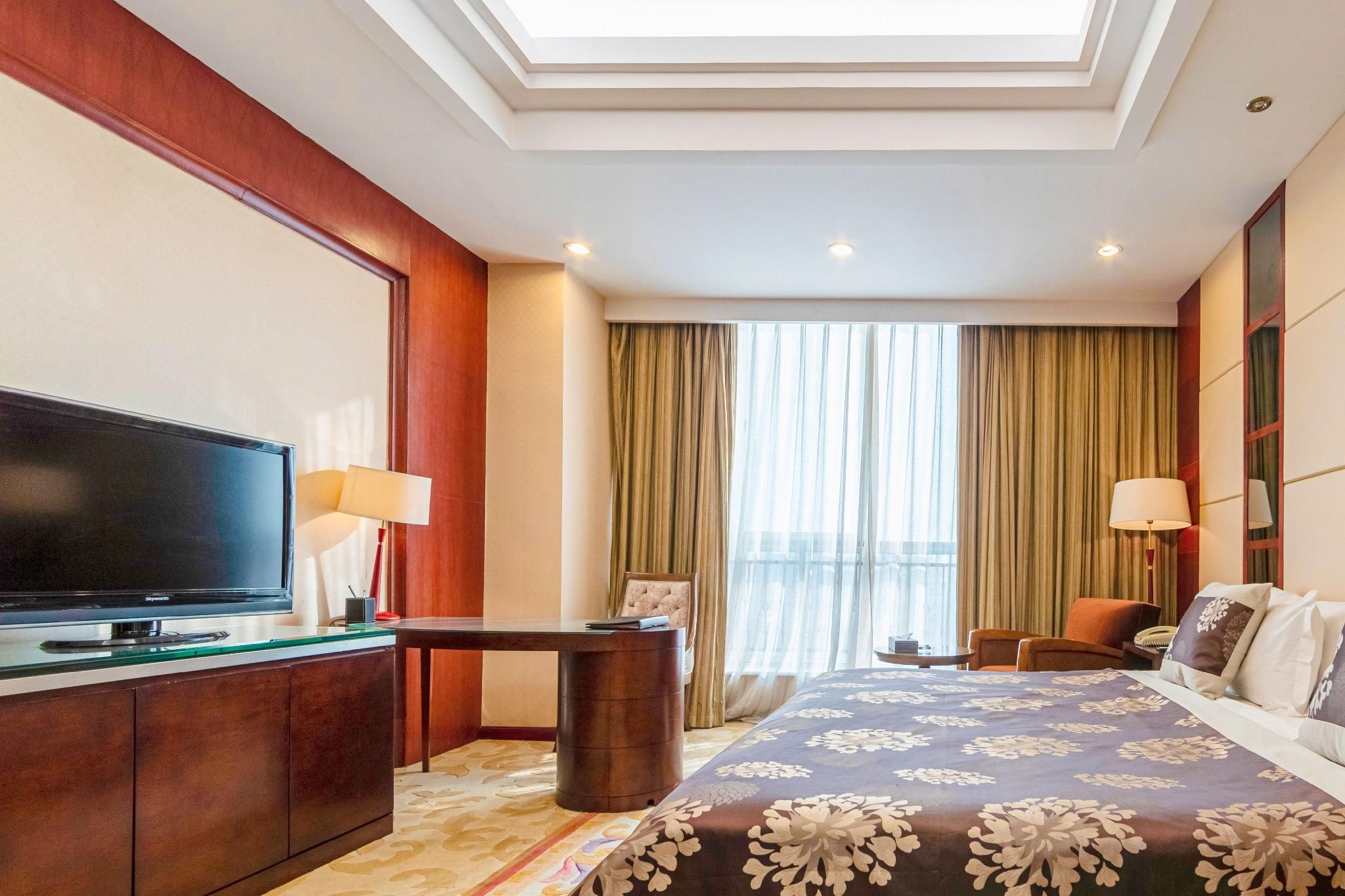 Habitación de hotel con televisor de pantalla plana, escritorio, cama con cubrecama floral, una silla y vista de la ciudad a través de amplias ventanas.