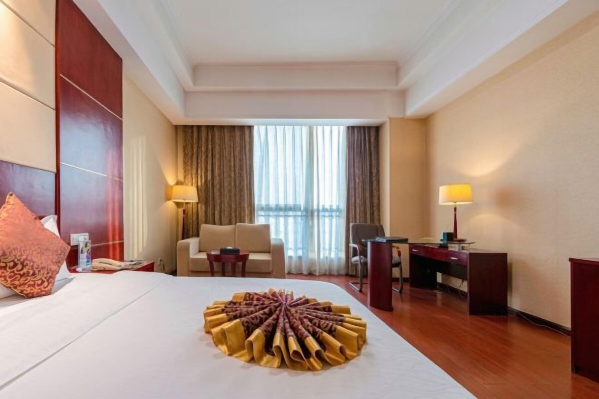 A hotel room with a neatly made bed, a desk, a chair, a sofa, and a large window with curtains.
