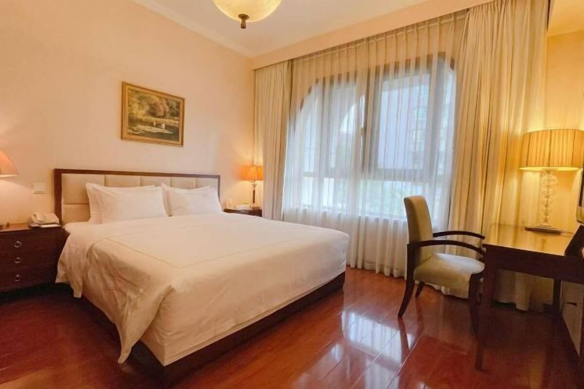 A hotel room with a bed, a desk, a chair, a lamp, a painting on the wall, and a window with curtains.