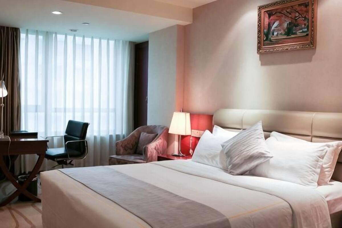 A hotel room with a bed, two chairs, a desk, a lamp, and a painting on the wall.