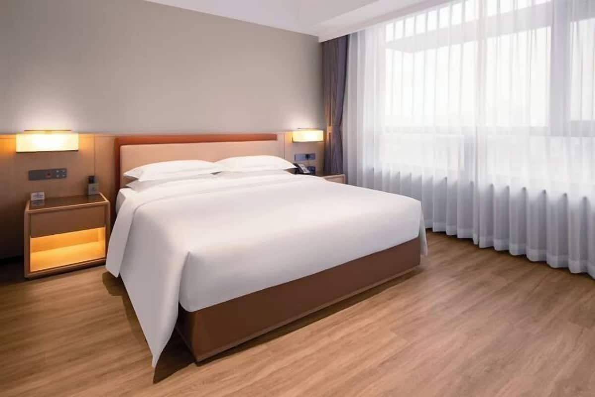 A hotel room with a large bed, bedside lamps, a nightstand, and a view of the outdoors through sheer curtains.