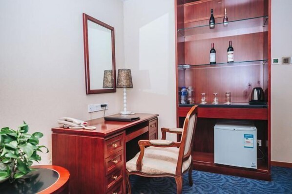 A hotel room with a wooden desk, a chair, a small refrigerator, a mirror, and a shelf with bottles and glasses. - Daxing Hotel (BEIJING)