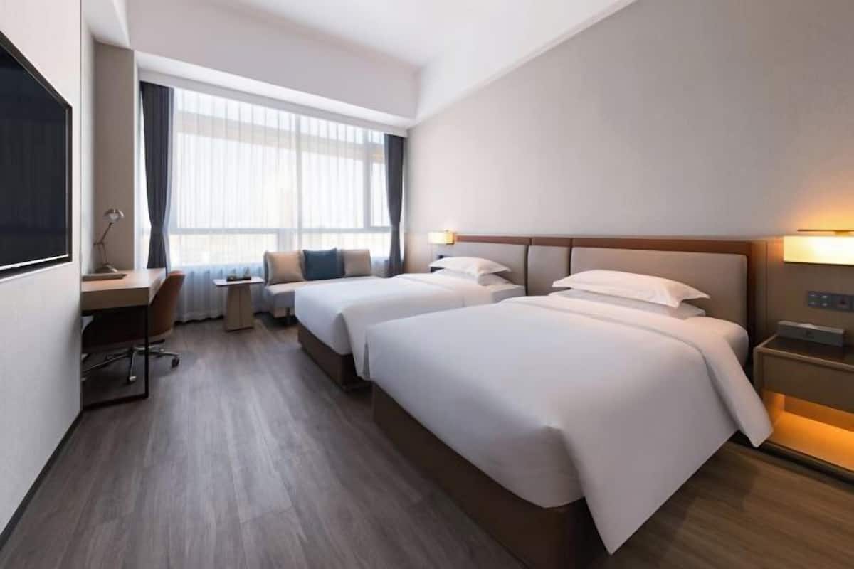 A hotel room with two beds, a desk, a chair, and a large window with curtains.