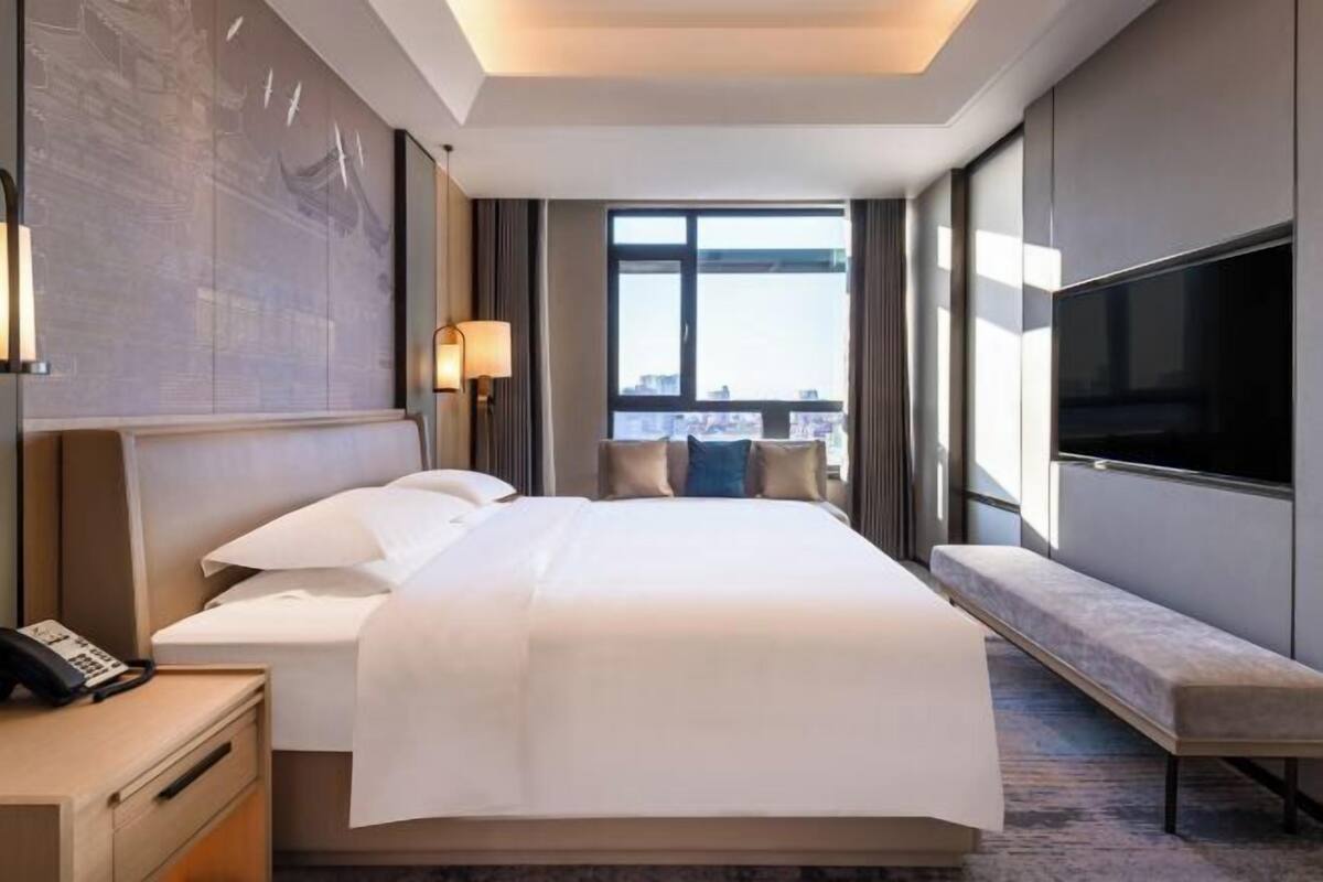 A modern hotel room with a large bed, a TV, a bedside table with a phone, and a view of the city through a large window.