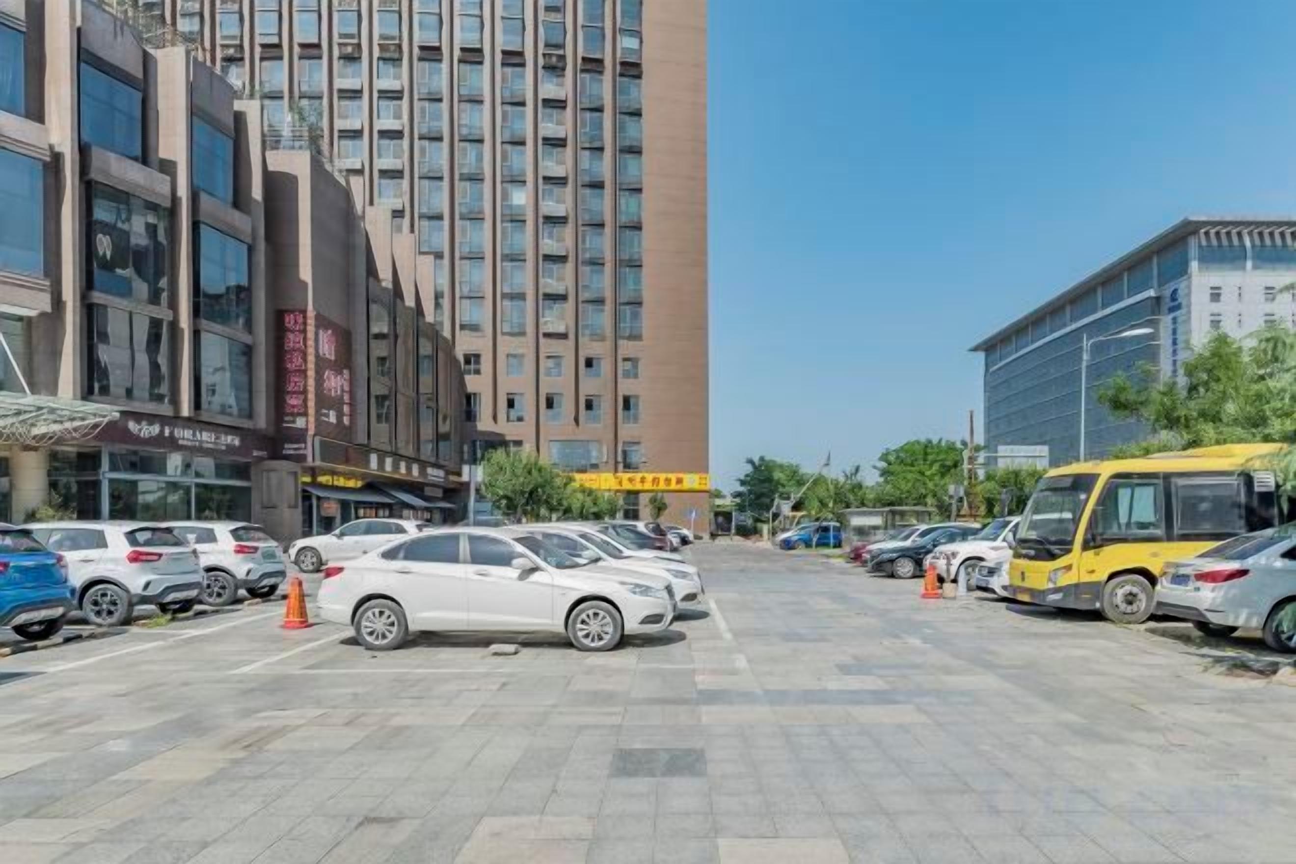 A parking lot with multiple cars and a yellow bus, surrounded by buildings with commercial signage.