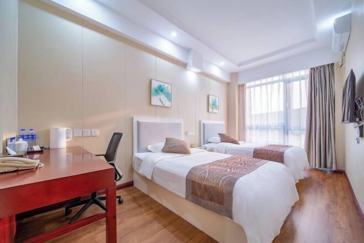 Habitación de hotel con dos camas, un escritorio y una silla. Hay cuadros en la pared, una ventana con cortinas y un equipo de aire acondicionado.
