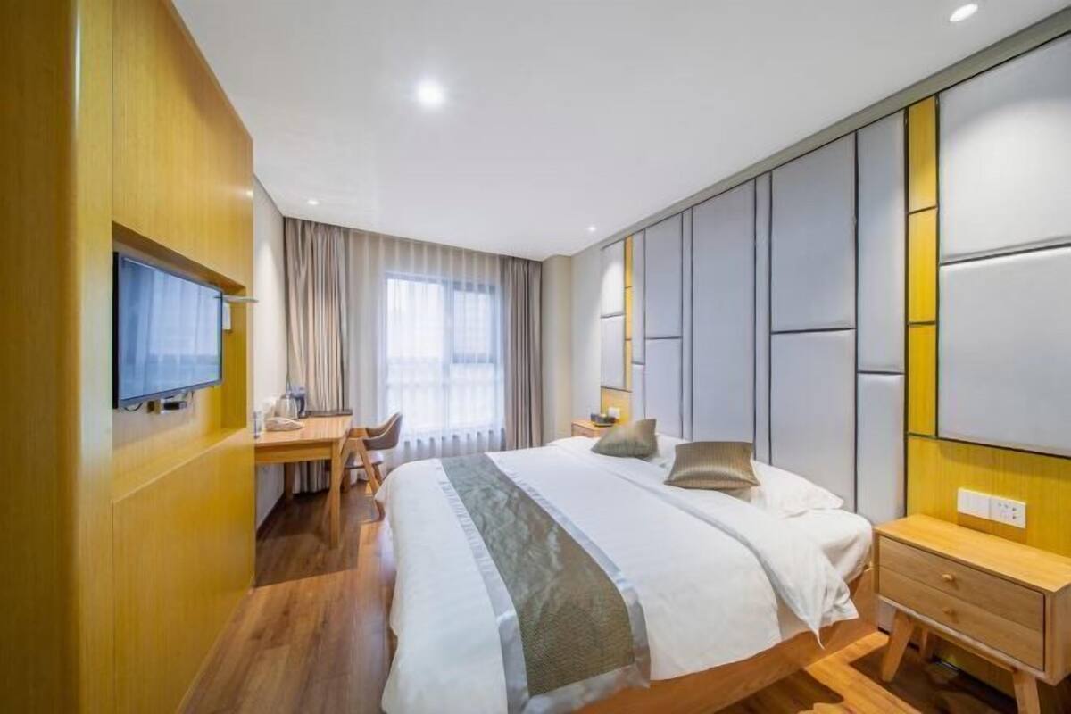 Habitación de hotel con cama, mesita de noche, televisión, escritorio, silla y un ventanal grande con cortinas.