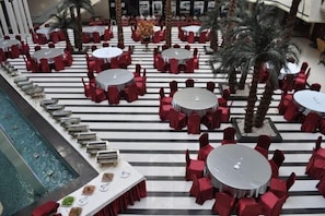 Vestíbulo de hotel con mesas redondas y sillas rojas, zona de piscina y palmeras.