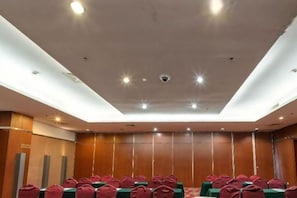 Een vergaderzaal met rijen tafels en stoelen, een hoog plafond met inbouwverlichting en een wand van houten panelen.