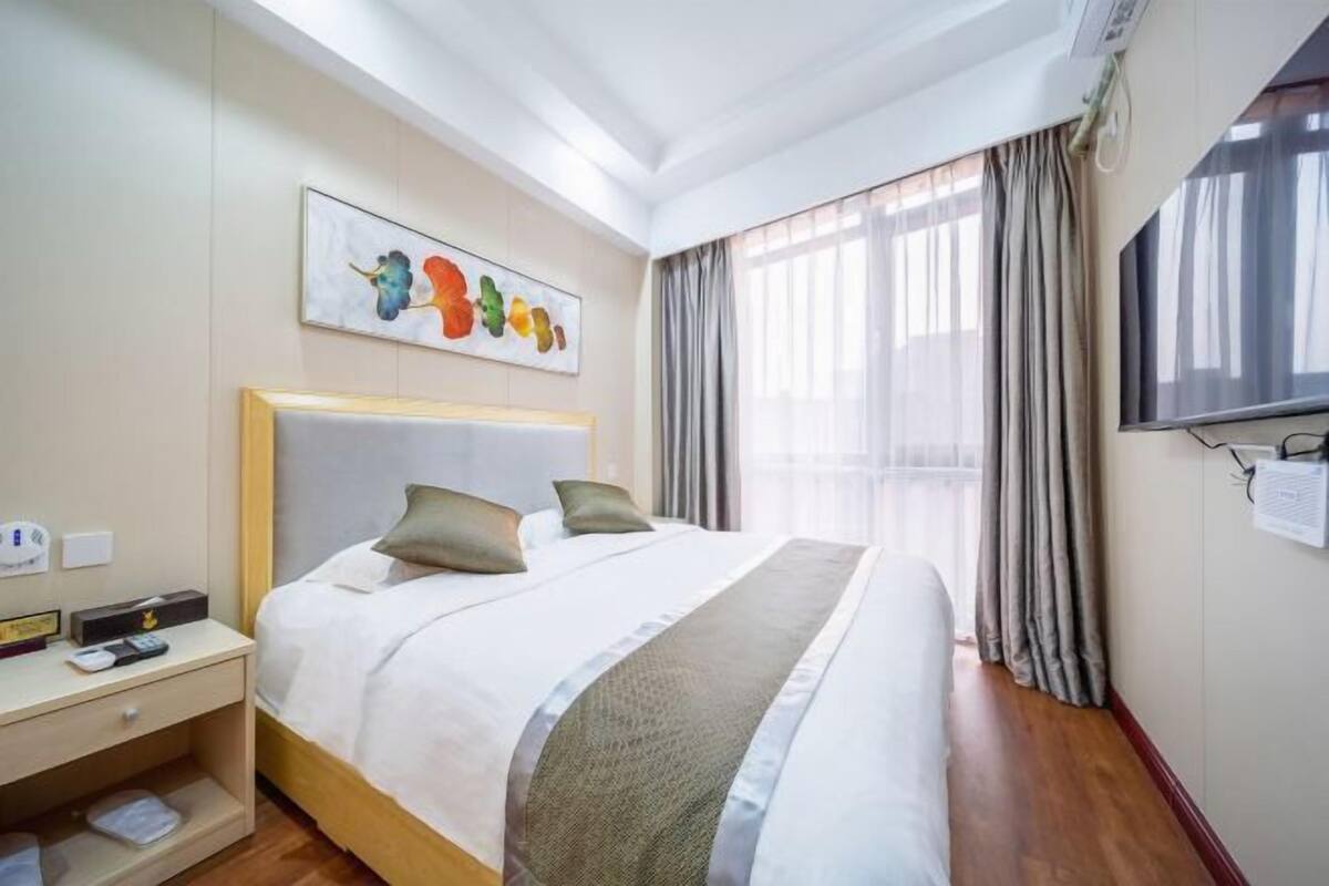 Habitación de hotel con cama, mesita de noche, televisor montado en la pared y ventana con cortinas.