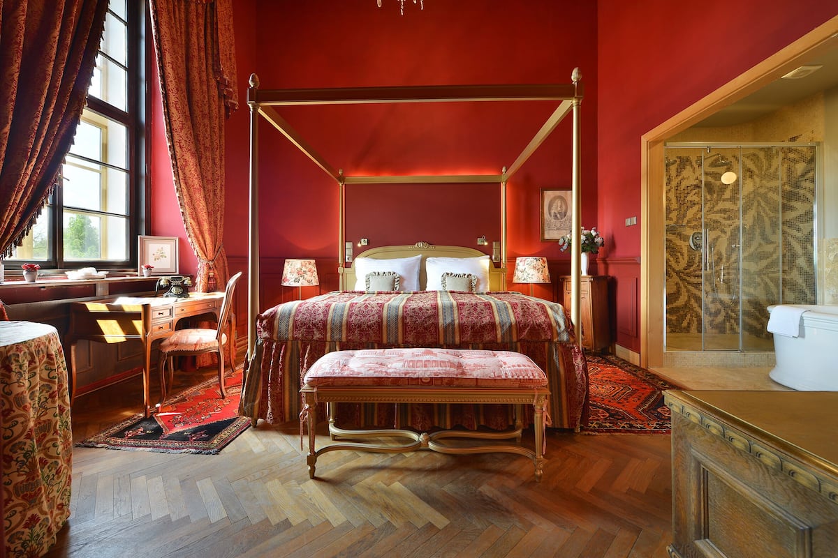 Een hemelbed met rode baldakijn, een bureau met stoel en een groot raam met gordijnen.