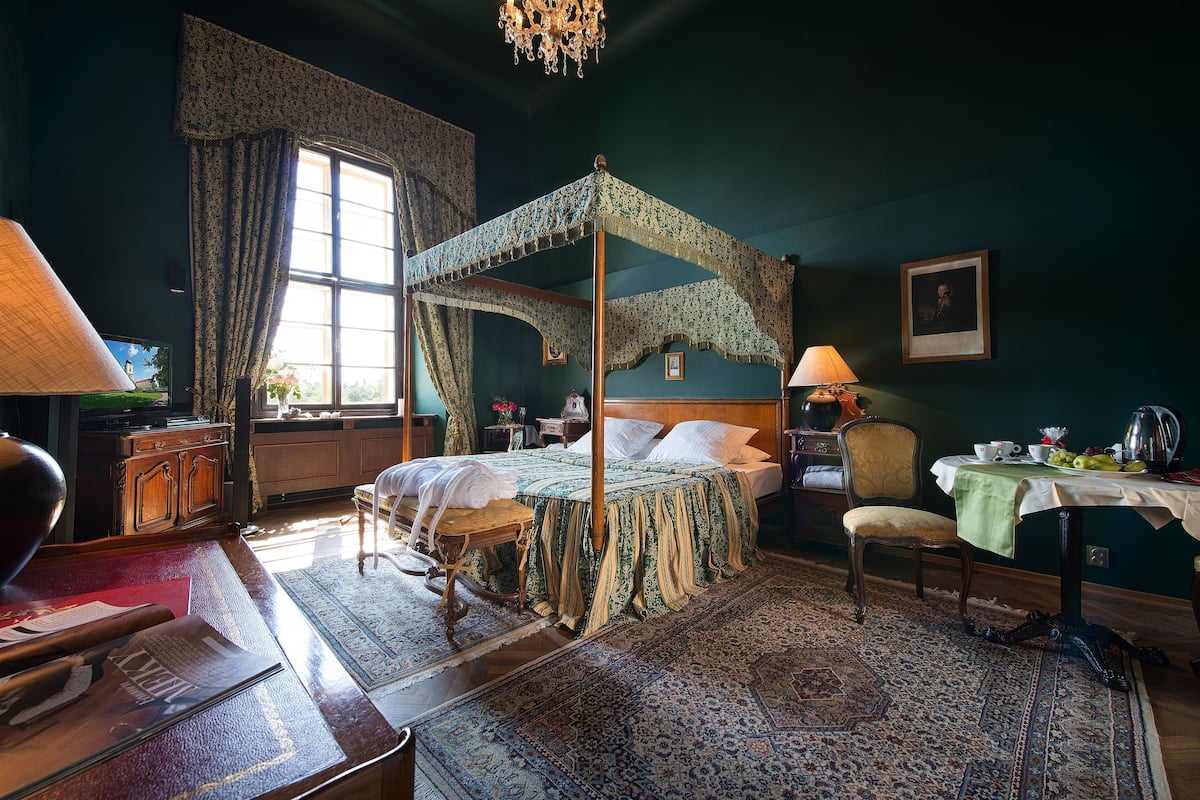 Een hemelbed met een groen beddegoed, een houten nachtkastje, een stoel, een tafel met een groene tafelkleed, en een kroonluchter.
