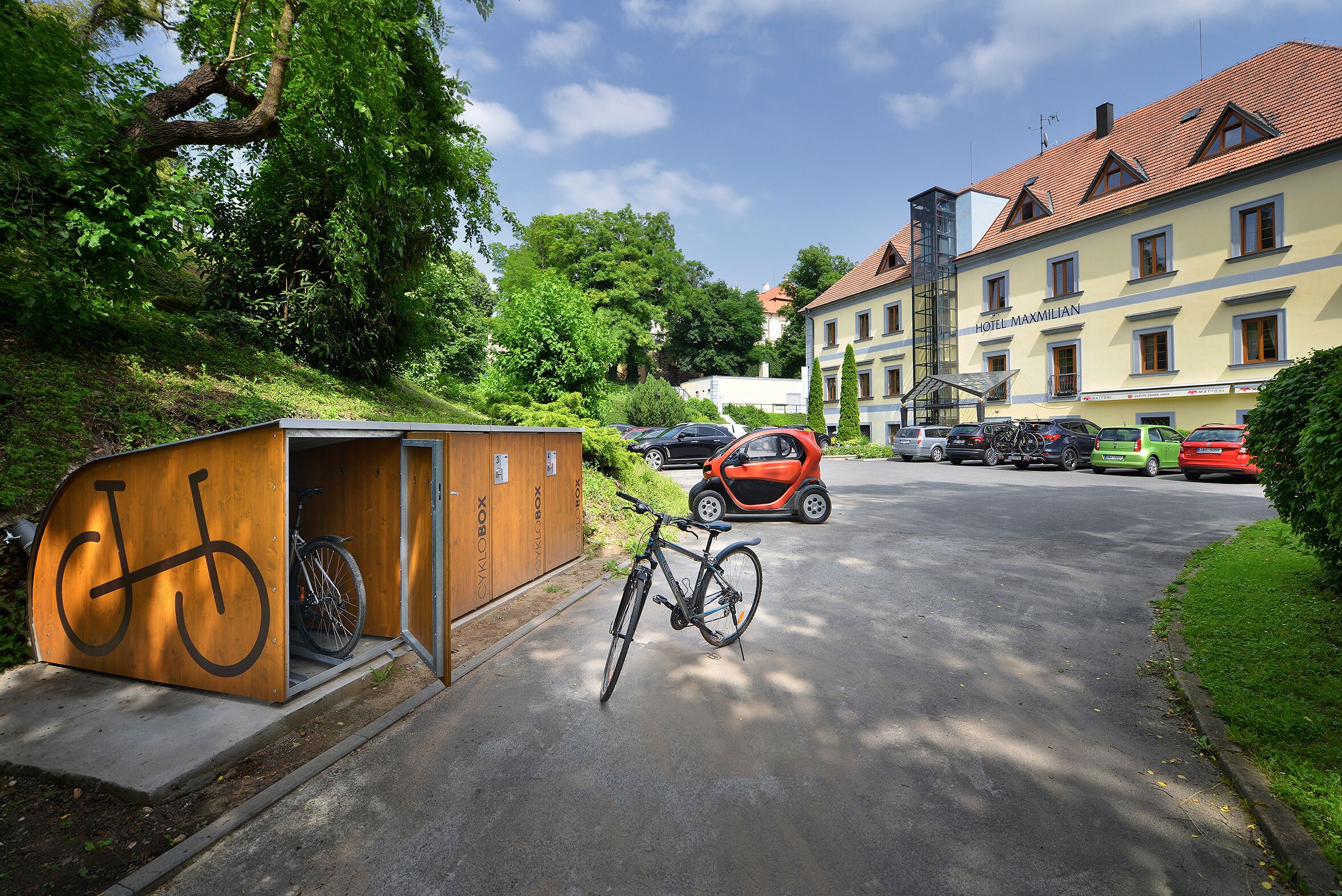 Un espacio para guardar bicicletas con una bicicleta dentro, un pequeño edificio con el símbolo de una bicicleta y un coche estacionado frente a un edificio con el letrero "HOTEL MAXMILIAN".