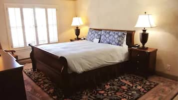 Un dormitorio con una cama grande, dos mesitas de noche con lámparas, una ventana con persianas y una alfombra estampada con motivos florales.