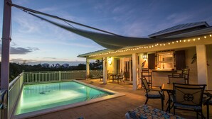 A poolside area with a covered seating area, a dining table with chairs, and a kitchen visible through an open door. - Casa de Isle (Providenciales)