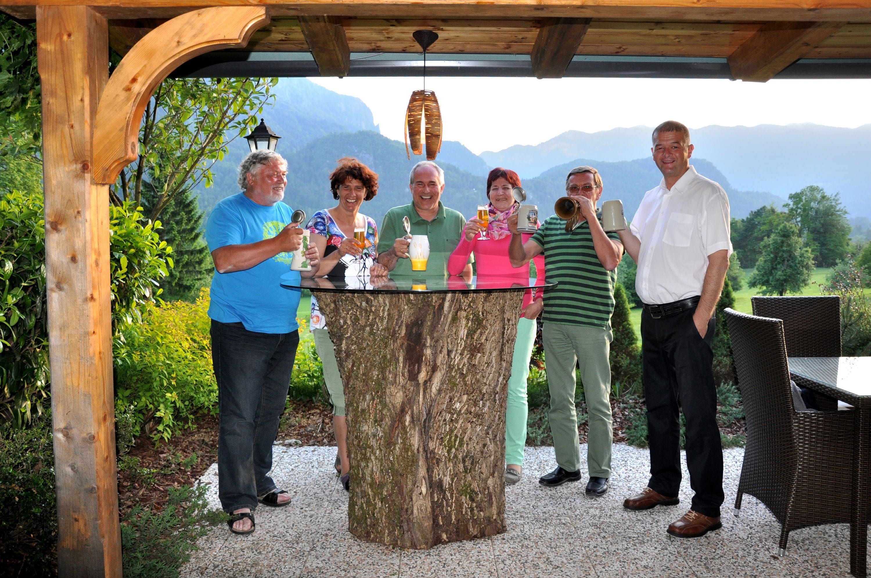 A group of people standing around a wooden table with a glass top, outdoors with a mountainous backdrop.