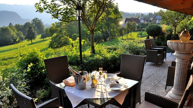 An outdoor dining area with wicker chairs and a table set for two, overlooking a lush green landscape with a view of houses in the distance. - Nature Hotel Lukanc Bled (Bled)