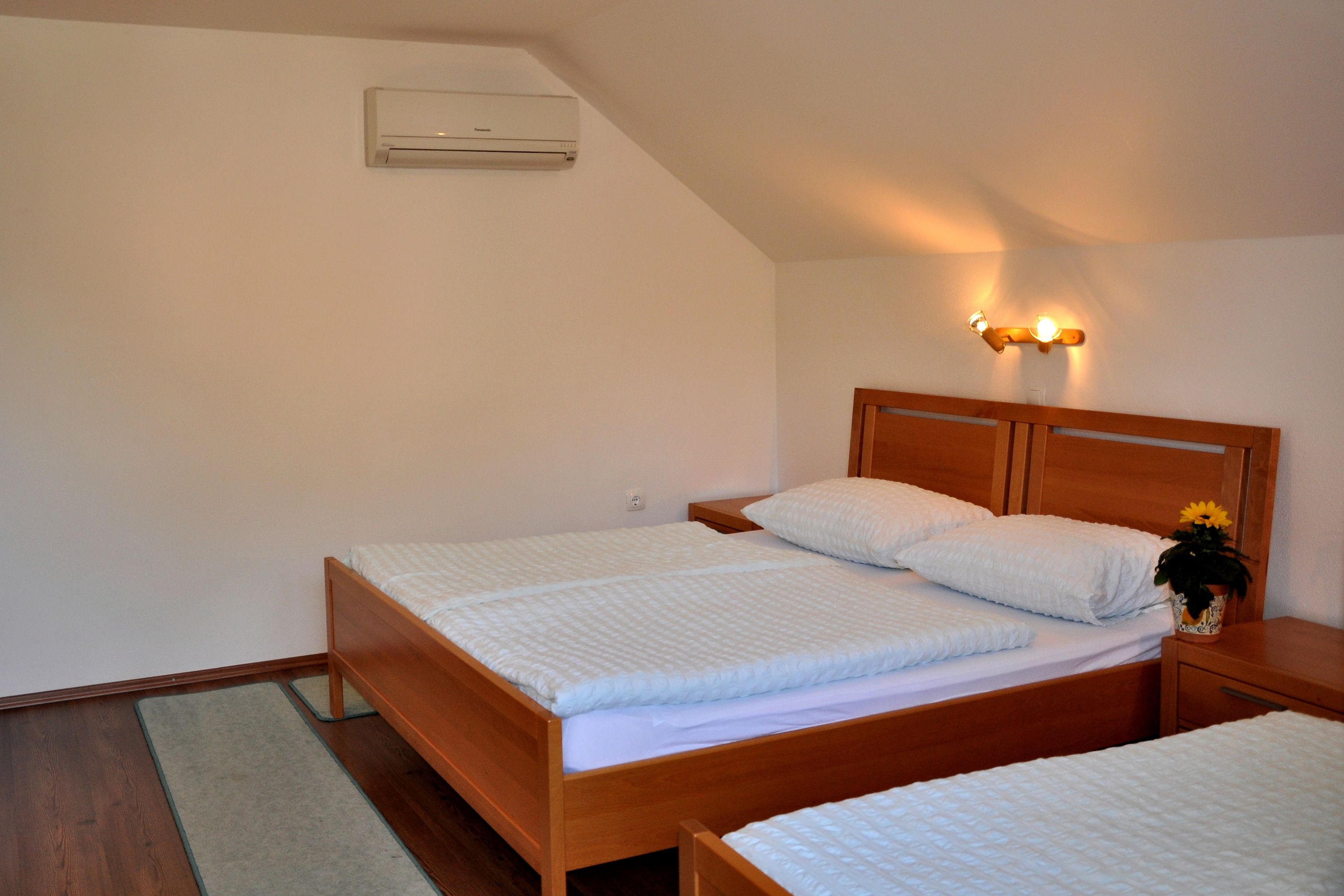 Une chambre avec deux lits simples, une tête de lit en bois, une table de chevet ornée d’une fleur et un climatiseur fixé au mur.