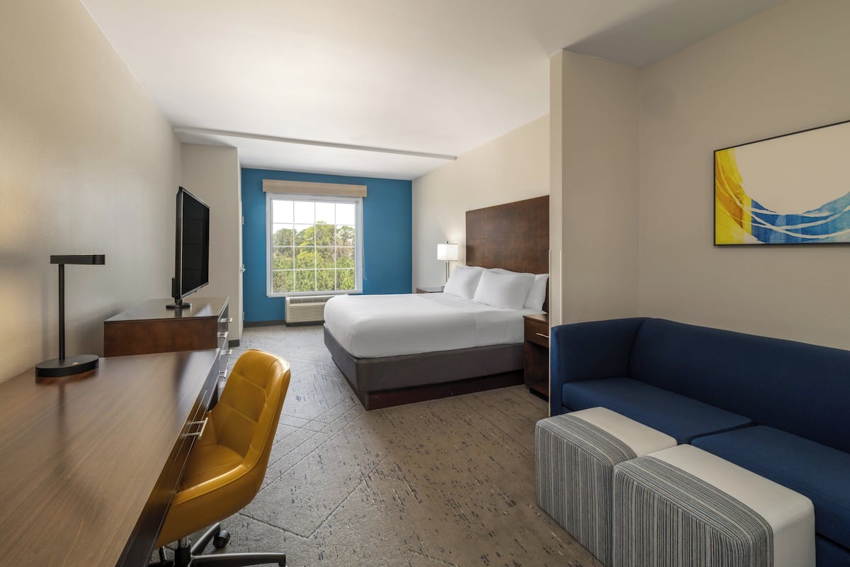 A hotel room with a bed, a desk with a chair, a sofa, a TV, and a window with a view of trees.