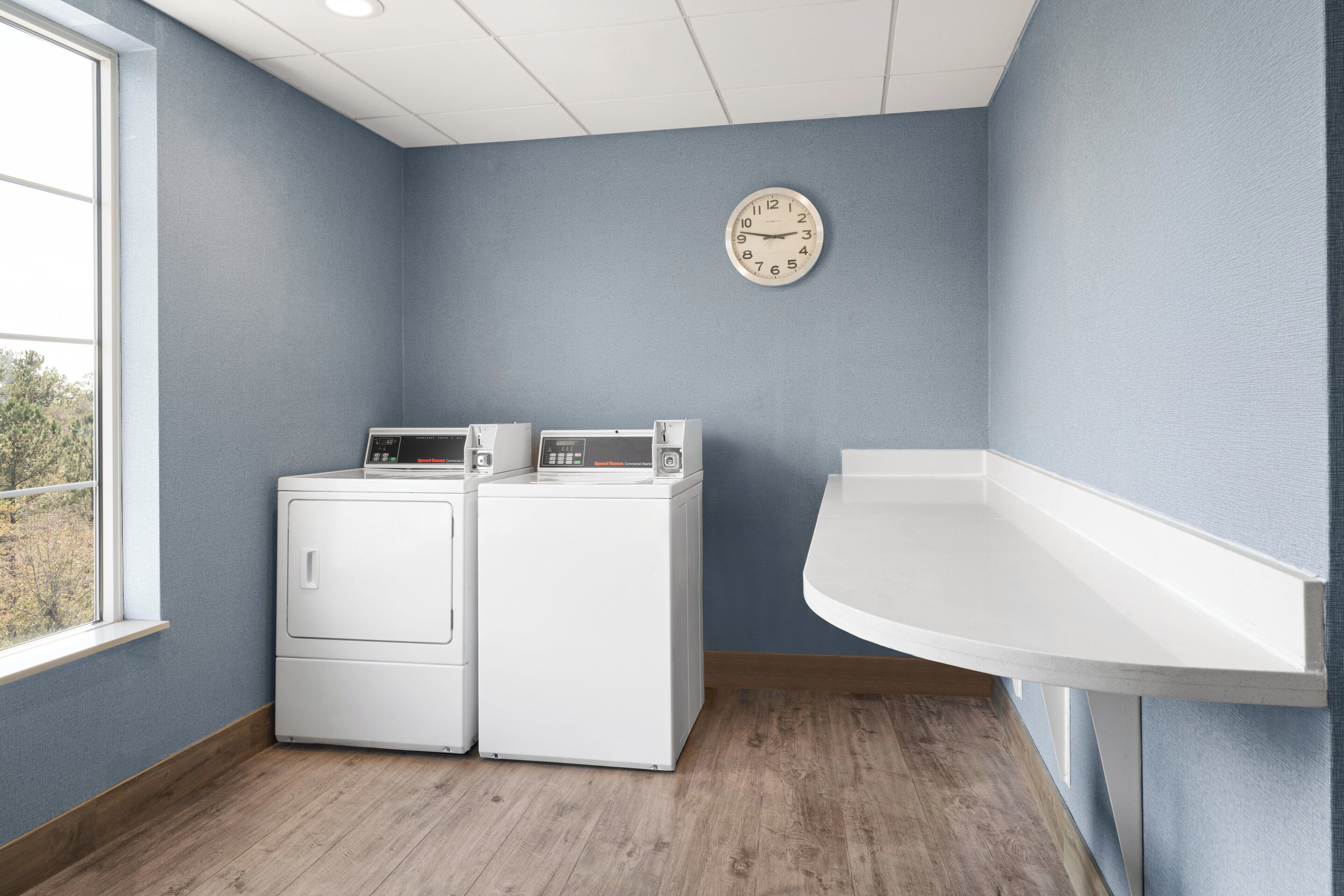 A laundry room with two white washers and dryers, a clock on the wall, and a window providing natural light.