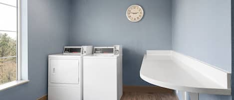 A laundry room with two white washers and dryers, a clock on the wall, and a window providing natural light.