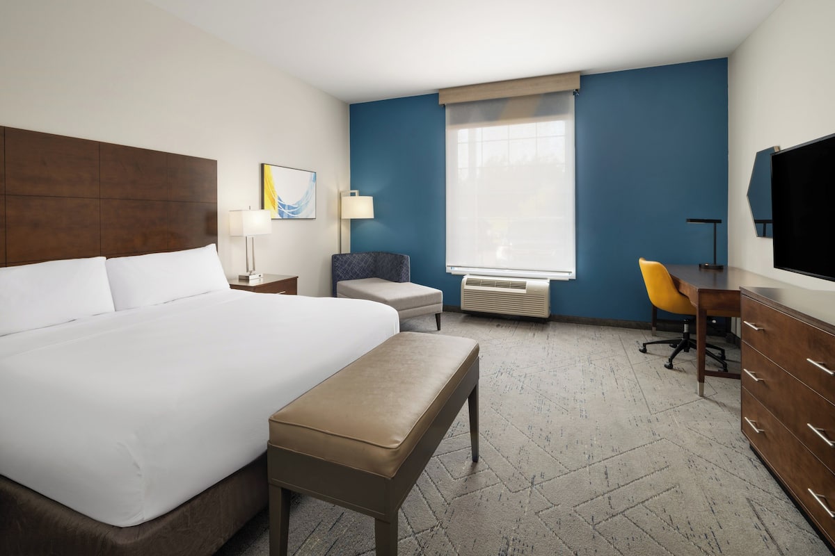 A hotel room with a large bed, a desk, a chair, a bench, a TV, and a window with curtains.