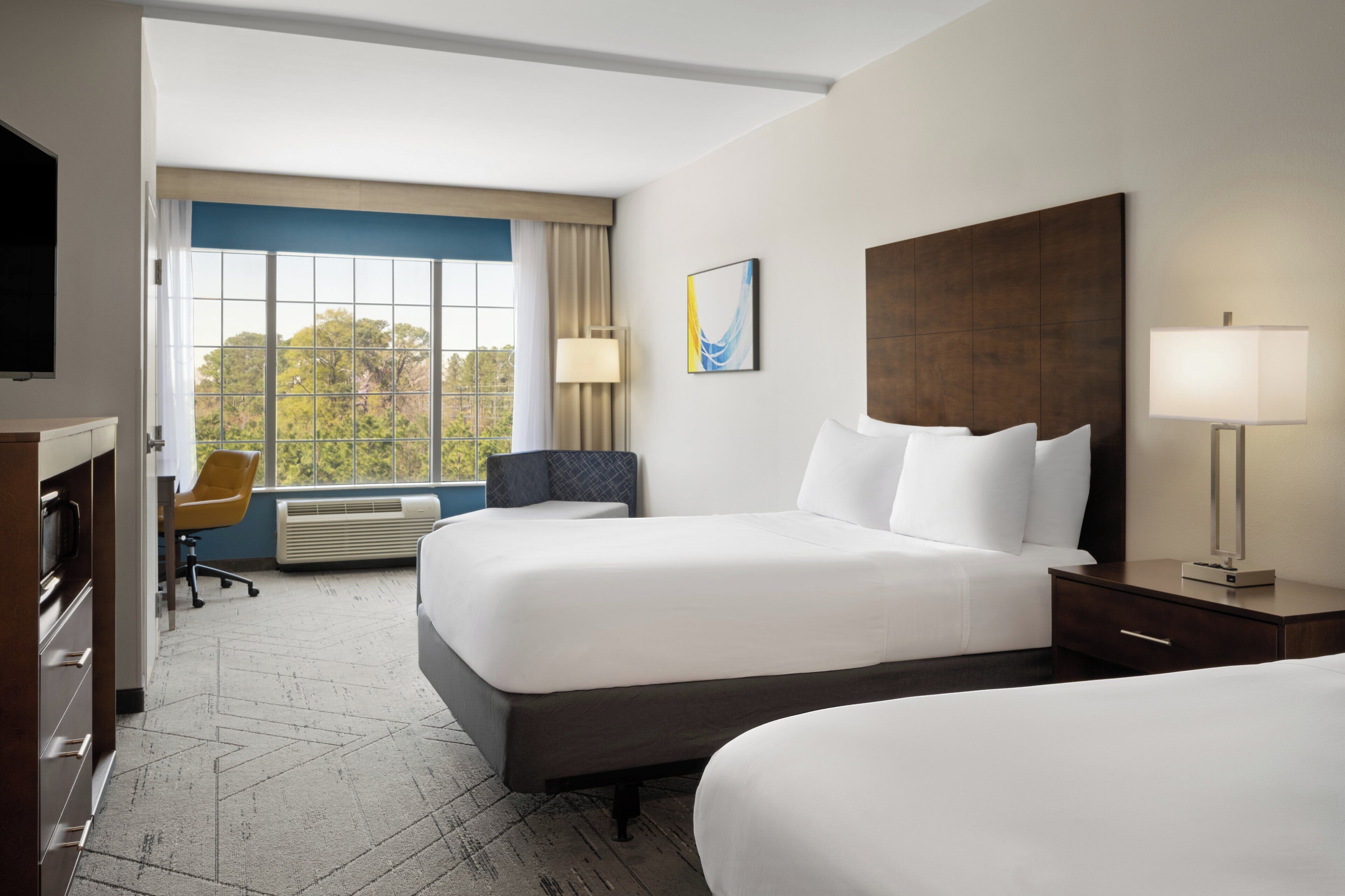 A hotel room with two beds, a desk, a chair, and a large window with a view of trees.