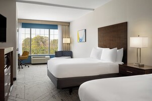 Habitación de hotel con dos camas, un escritorio, una silla y un ventanal con vistas a árboles.