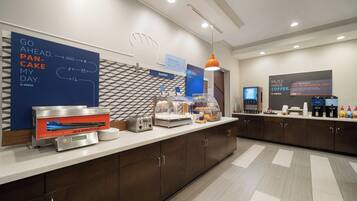 A modern breakfast area with a flat-screen TV, a coffee station, and a sign promoting the breakfast menu.
