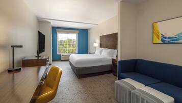 Habitación de hotel con cama, escritorio con silla, sofá, televisor y ventana con vista a árboles.