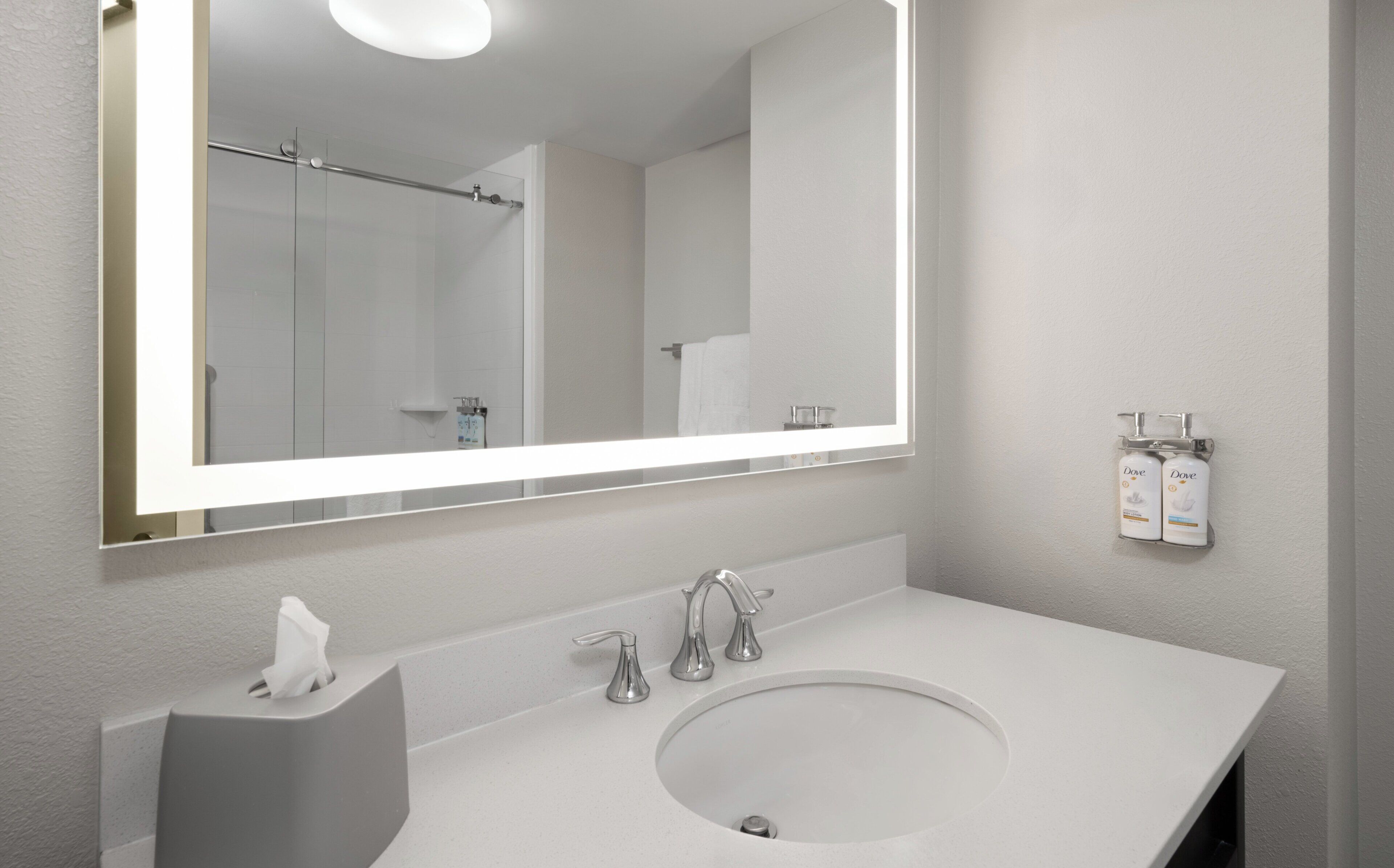 Un baño moderno con un espejo grande, un lavamanos blanco y dos dispensadores de jabón Dove en la pared.