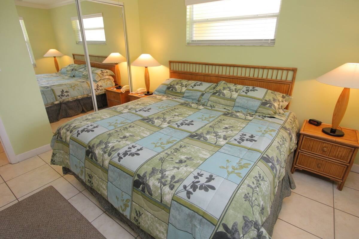 A bedroom with a large bed, two bedside tables, a nightstand, a mirror, and a window with blinds.
