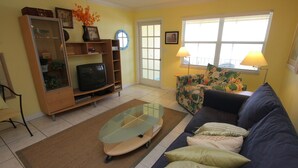 Ein Wohnzimmer mit einer Couch, einem Glastisch, einem Fernseher, einem Bücherregal und einem Fenster mit Jalousien.