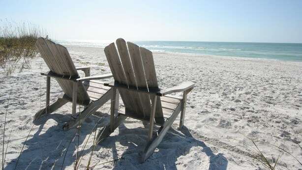 Zwei hölzerne Adirondack-Stühle an einem sandigen Strand mit Blick auf den Ozean.