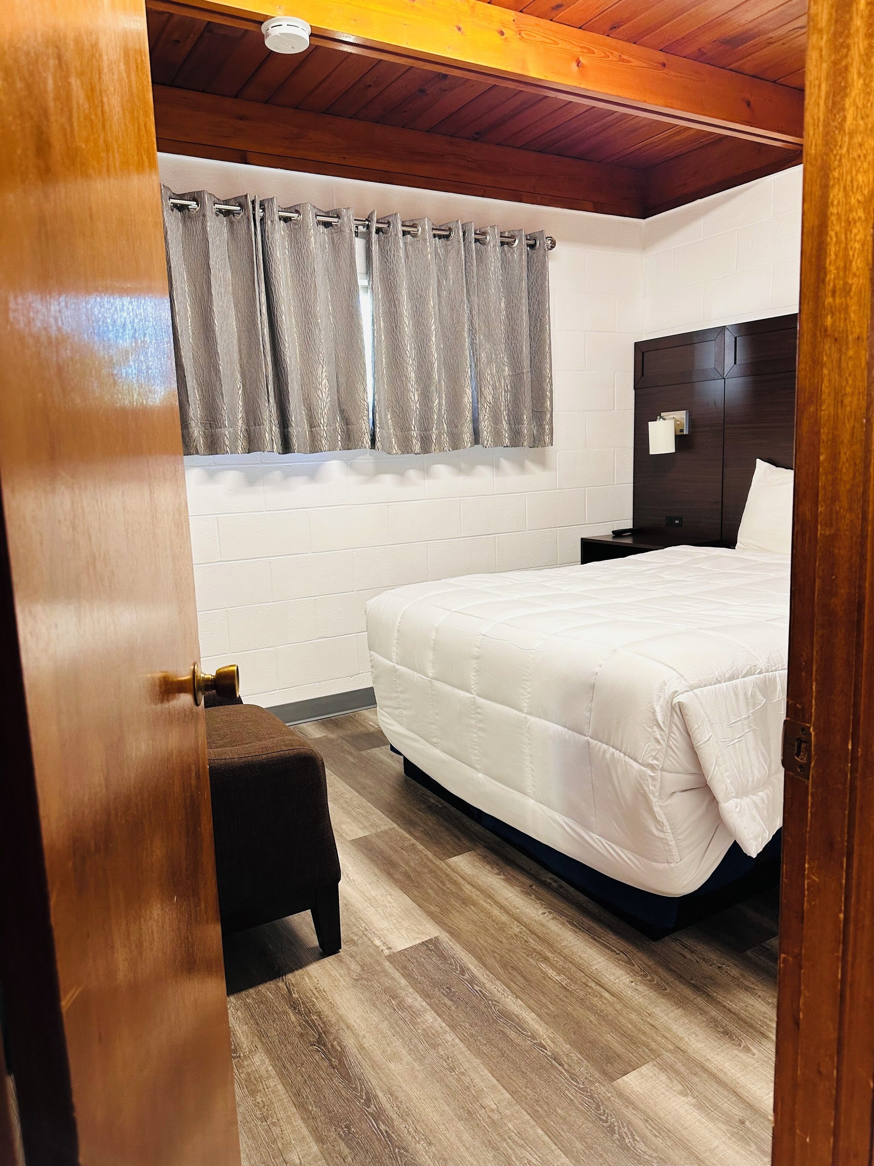 A bedroom with a wooden ceiling, a bed with a white mattress, a nightstand, a lamp, and a window with curtains.