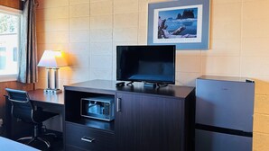 Ein kleines Hotelzimmer mit Holzboden, einem Fernseher auf einer Holdeklage, einer Mikrowelle, einem Kühlschrank, einem Schreibtisch mit Stuhl und einem Fenster mit Vorhängen.