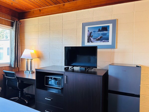 Habitación de hotel compacta con techo de madera, televisor sobre un mueble de madera, microondas, refrigerador, escritorio con silla y ventana con cortinas.